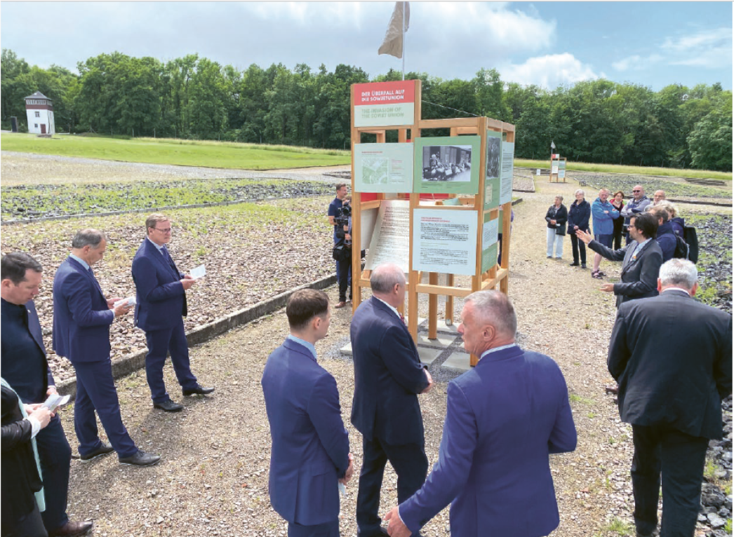 Mehrere Personen stehen im Freien um eine hölzerne Ausstellungsstruktur mit Informationstafeln zur Ausstellung „Der Überfall auf die Sowjetunion“. Einige Besucher:innen hören einem sprechenden Mann zu, andere lesen die Texte. Die Installation befindet sich auf dem Gelände der Gedenkstätte, umgeben von offenen Flächen und Bäumen.
