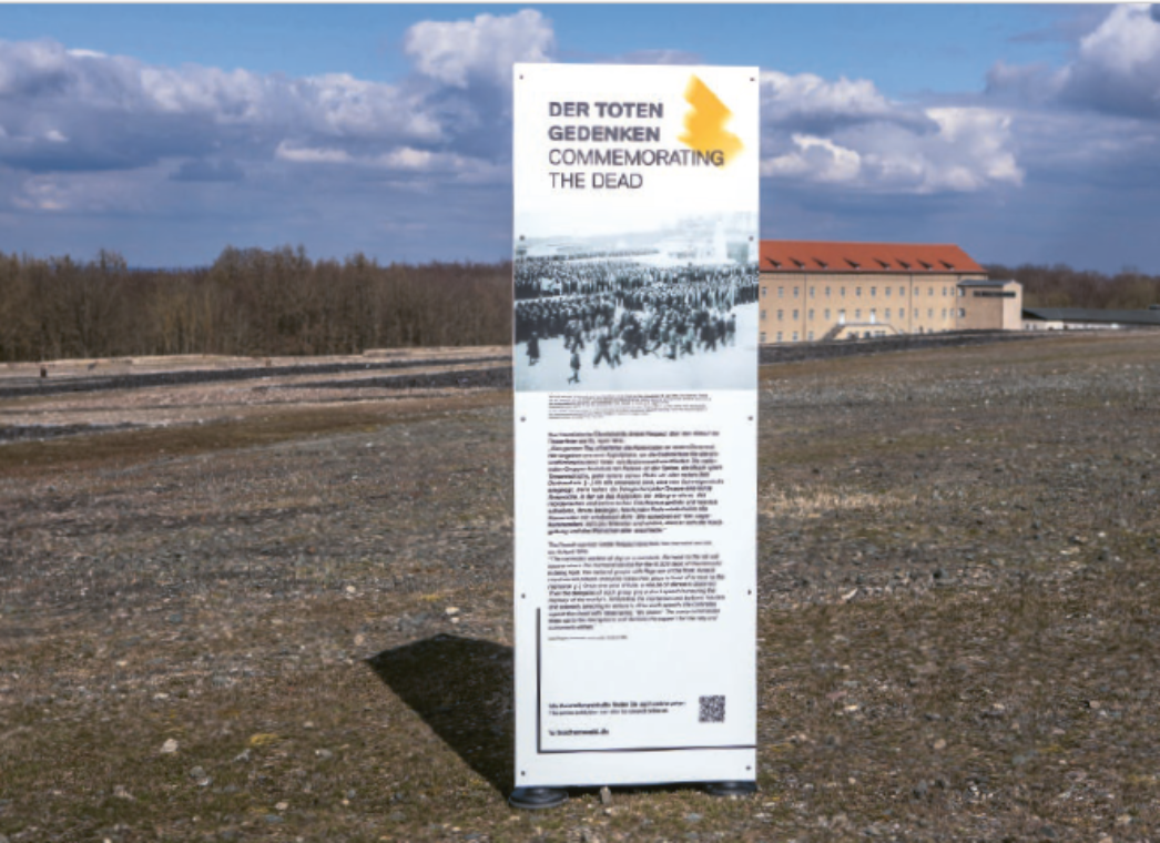 Freistehende Stele mit der Überschrift „Der Toten gedenken / Commemorating the Dead“ auf dem ehemaligen Appellplatz des KZ Buchenwald. Auf der Tafel sind ein historisches Foto sowie erklärende Texte zu sehen. Die Stele steht in einer weiten, offenen Landschaft; im Hintergrund ist ein Gebäude der Gedenkstätte sichtbar.