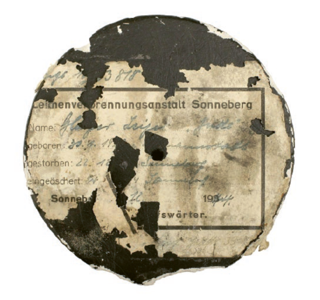 Runder, stark beschädigter Papieraufkleber mit Brandspuren und fehlenden Teilen. Der Aufkleber stammt von einer Urne und trägt die Aufschrift „Leichenverbrennungsanstalt Sonneberg“ sowie handschriftlich eingetragene Angaben zu einer verstorbenen Person.