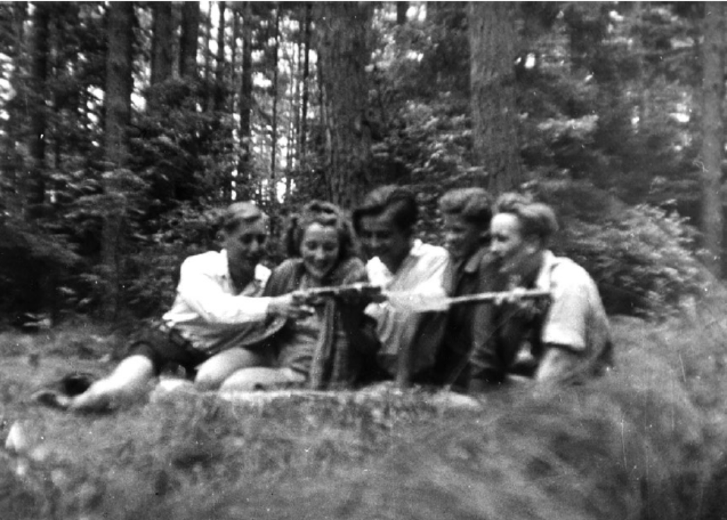 Historisches Schwarzweißfoto von fünf Jugendlichen, die im Wald auf dem Boden liegen. Sie lachen und halten gemeinsam Stöcke oder Gegenstände in den Händen, während sie eng beieinanderliegen und in die gleiche Richtung schauen.