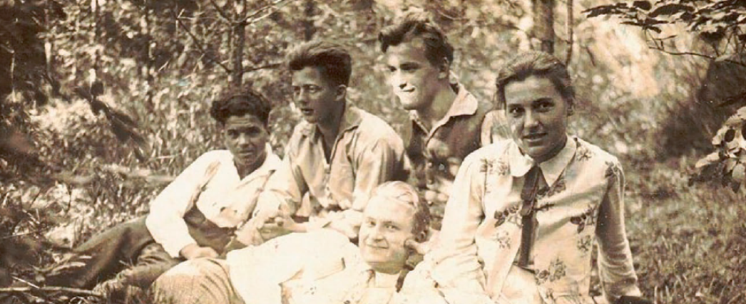 Historisches Schwarzweißfoto von fünf jungen Menschen, die entspannt im Freien auf dem Boden sitzen bzw. liegen. Sie befinden sich in einer bewaldeten Umgebung, schauen teilweise in die Kamera und wirken wie eine Gruppe von Freund:innen oder Bekannten.