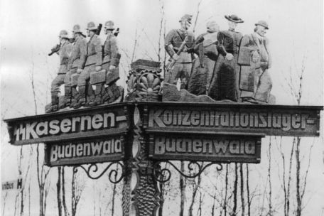  A hand-carved signpost to the Buchenwald SS barracks and Buchenwald concentration camp on Carachoweg. Above the lettering you can see figures carved out of wood. Left: SS soldiers, right: an SS man driving three people in front of him. The figures are depicted as a monk, priest, and man with suit and hat.