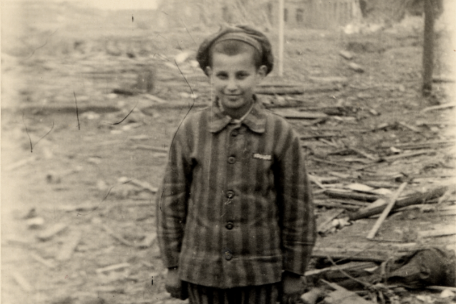A boy in prisoner's clothing stands in front of rubble and smiles at the camera.