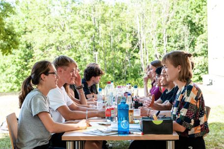 Zu sehen sind mehrere Jugendliche an einem Tisch in der Nähe der Internationalen Bildungsstätte, die angeregt miteinander diskutieren. Neben Getränken ist Lehrmaterialauf den Tischen verstreut.