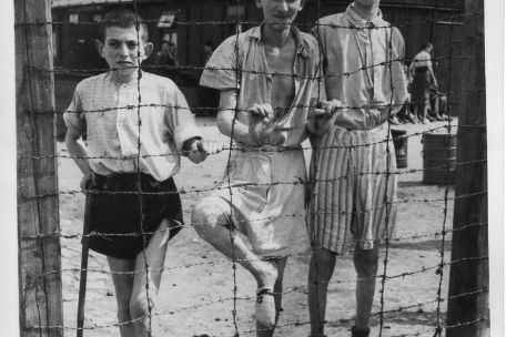 Three liberated youths at the barbed wire fence, in the middle Abram Zylbersztajn. One is leaning on a stick. Behind them a wooden barrack and two stone blocks