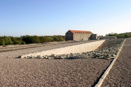 On the gravel ground of the camp area the areas of the former barracks are marked with dark stones. The surface of one barrack is densely filled with larger pieces of stone. Towards the center, the surface slopes significantly, resulting in a stone depression.