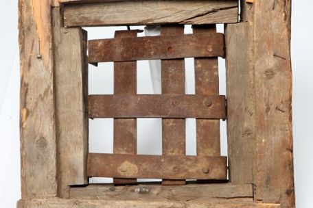 A wooden door made of rough beams. A window in it is barred with wide iron bars. 