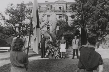 In the foreground of the picture you can see two people with their backs to the photographer. They are wearing large frames. Further back in the picture, 3 people are standing next to a wreath laid on the ground.