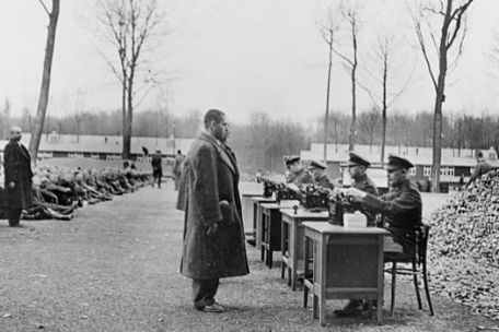 On the right side of the road, there are four tables outside at which uniformed people are sitting. People in civilian clothes are standing in front of the tables. On the left side of the path, numerous other people are sitting on the ground. In the background, flat functional buildings.