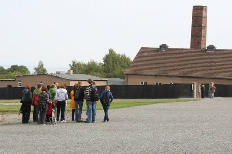 View over the roll call square. In the background the former crematorium with a high chimney. A small group of visitors is standing on the square. 