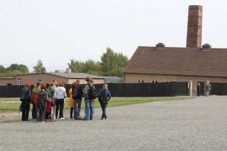 Blick über den Appellplatz. Im Hintergrund das ehemalige Krematorium mit einem hohen Schornstein. Auf dem Platz steht eine kleine Gruppe Besucher:innen. 