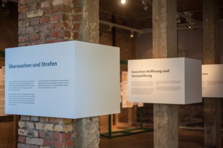 Säulen aus Ziegelsteinen und Holz tragen Kästen, die Informationstexte zur Ausstellung zeigen. 