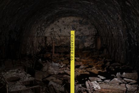 Eine gelbe Stele mit der Aufschrift "Chamber 46" steht im Stollen. Im Hintergrund sind am Boden Trümmerteile der Stollenanlage zu erkennen.