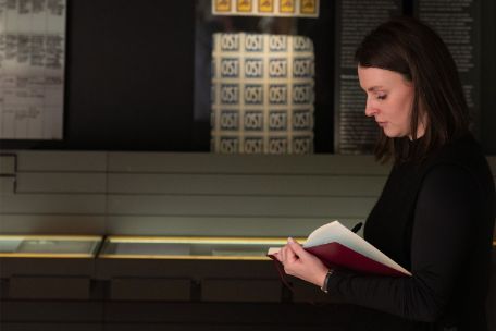 Eine Frau steht in einer Ausstellung und liest in einem roten Notizbuch. Im Hintergrund sind Vitrinen und Ausstellungstafeln mit historischen Dokumenten und Objekten zu sehen. Das Foto verbildlicht Bildungsreisen.