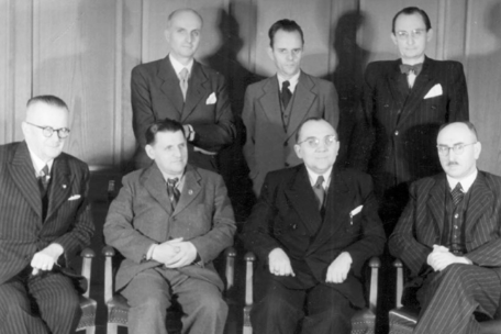 Schwarz-weiß-Gruppenporträt vom 5. Dezember 1946: Sieben ehemalige Buchenwald-Häftlinge in formeller Kleidung als Mitglieder der Thüringer Landesregierung. In der vorderen Reihe sitzen unter anderem Ministerpräsident Rudolf Paul (3. von links) sowie Innenminister und stellvertretender Ministerpräsident Ernst Busse (2. von links). Hinter ihnen stehen weitere Regierungsmitglieder, darunter Walter Wolf (2. von links), Volksbildungsminister. Die Aufnahme zeigt die frühe Nachkriegsregierung in offizieller Pose.