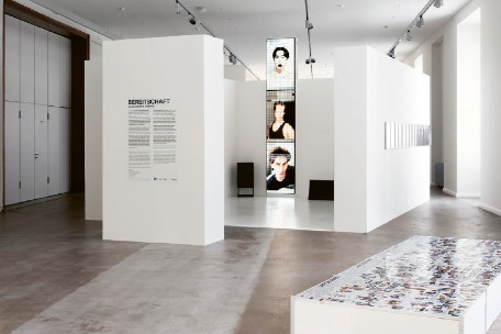 Ausstellungsraum eines Museums mit weißen Stellwänden und klarer, moderner Gestaltung. Auf einer Wand ist der Titel „Bereitschaft“ mit Begleittext zu sehen. In der Mitte steht eine schmale, hohe Videoinstallation mit mehreren Bildsequenzen von Gesichtern. Im Vordergrund befindet sich eine niedrige Vitrine oder Plattform mit vielen ausgelegten Bildern oder Dokumenten.