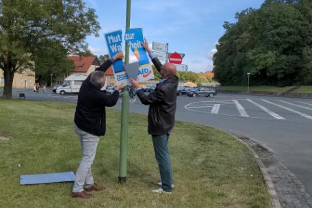 Two men remove an AfD poster from a lamppost at the memorial parking lot. It bears the slogan "Courage for truth