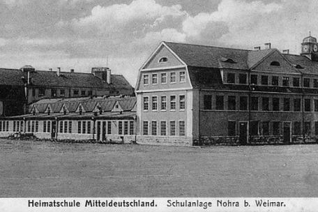  Ausschnitt aus einer historischen Postkarte. Sie zeigt ein großes Gebäude mit zwei höheren Flügeln, die in der Mitte durch einen flachen Teil verbunden sind. Darunter steht "Heimatschule Mitteldeutschland. Schulanlage Nohra bei Weimar"