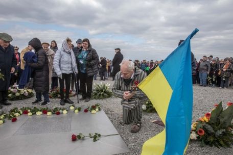 An elderly man in prisoner's clothing kneels in front of the memorial slab with flowers laid on its edges. He holds a Ukrainian flag. Around him numerous other people. 