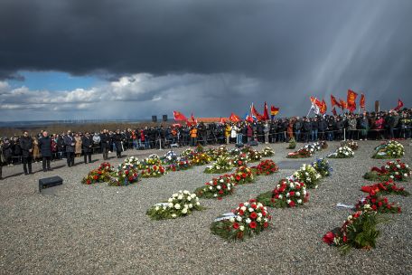 Auf dem Appellplatz sind Kränze in Reihen niedergelegt. Sie sind von einem Absperrband umgeben. Dahinter stehen zahlreiche Menschen, die teilweise Flaggen tragen. Dunkle Wolken sorgen für eine dramatische Beleuchtung. 