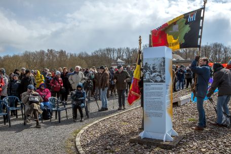 Im Vordergrund des Bildes steht eine weiß-grau-orangene Info-Stele. Rechts neben der Stele steht ein Mann mit einer großen Belgischen Flagge. Im Hintergrund ist eine Menschenmenge zu sehen, die in Richtung der Stele blickt. 