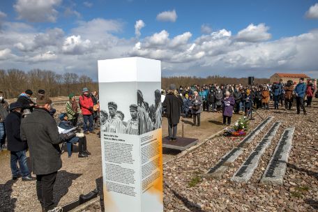 In the foreground there is a white-grey-orange information desk on which explanations are offered in various languages. Furthermore, on all sides are cutouts of a photograph of Burgenland Roma in prisoners' clothing. To the left of the stele is a speaker at a microphone. In the background, a crowd of people can be seen looking in the direction of the speaker.