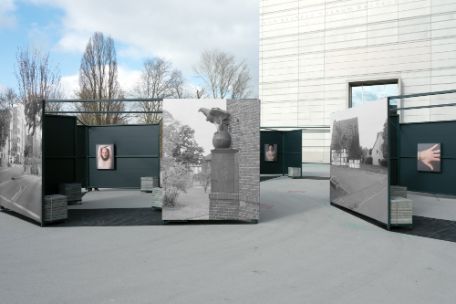 In a public square there are metal boxes with photos hanging on their walls. On the outsides are large photos of buildings in Weimar in the 40s, inside photos of people showing their tattoos. 