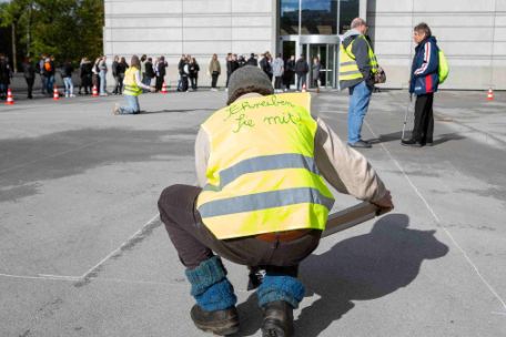 Ein Mann kniet auf einem Steinplatz und trägt eine gelbe Warnweste mit der Aufschrift "Schreiben Sie mit" auf dem Rücken. In den Händen hällt er ein Klembrett, welches er gerade vor sich legt. Im Hintergrund stehen und knien weitere Menschen mit Warnwesten auf dem Platz.