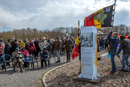 In the foreground of the picture there is a white-gray-orange info stele. To the right of the stele is a man with a large Belgian flag. In the background there is a crowd of people looking towards the stele. 
