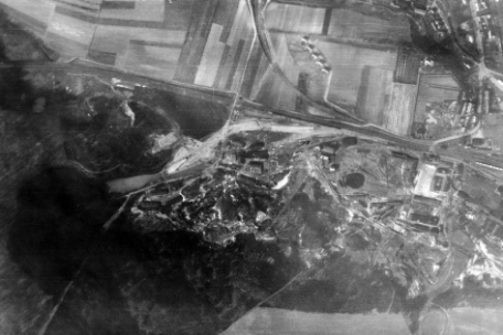 Schwarz-weiß-Luftaufnahme einer weitläufigen Landschaft mit Feldern, Straßen und einem größeren Lager- oder Industriekomplex. Mehrere Gebäude und Anlagen liegen am Rand eines Waldgebiets; Wege und Gleise verbinden das Gelände mit umliegenden Ortschaften.