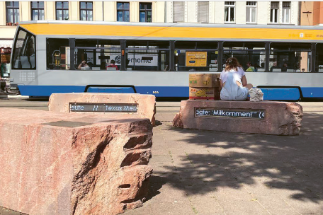 Drei große Steinblöcke im öffentlichen Raum, die als Gedenkzeichen gestaltet sind, stehen auf einem Platz. Auf ihnen befinden sich eingelassene Textbänder. Im Hintergrund fährt eine Straßenbahn vorbei, und eine Person sitzt auf einem der Steine.