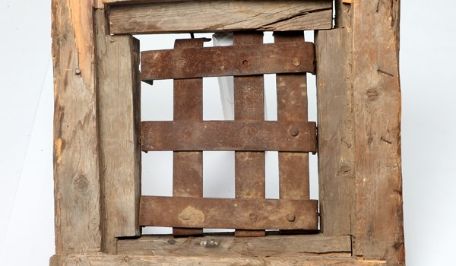 Eine Holztür aus groben Balken. Ein Fenster darin ist mit breiten Eisenstäben vergittert. 