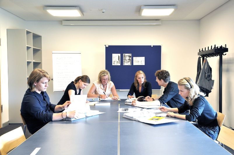 5 Junge Bundesfreiwiligendienstleistende sitzen an einem Tisch im Zentrum eines Seminarraumes und inspizieren die vor ihnen liegenden Papiere. Am Kopf des Tisches sitzt eine Frau die etwas älter aussieht.