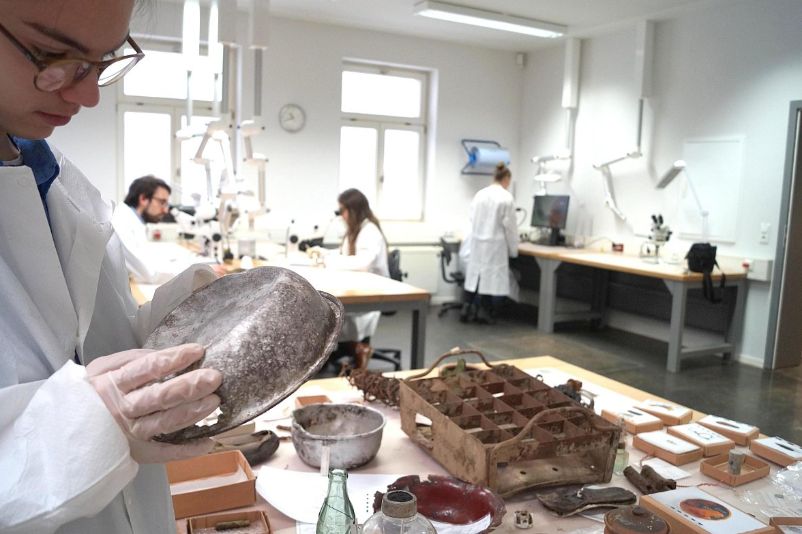 A person cleans an old eating bowl. On the table in front of the person are historical objects. People in lab coats in the background at equipment.