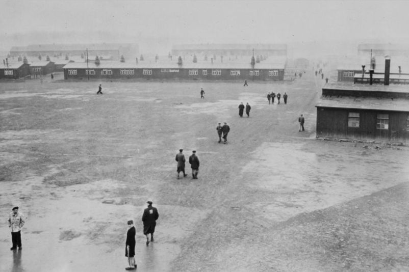 View from the tower of the gate building over the roll call area and the barracks in northern direction. The rear area of the camp disappears in the fog. In the large open area, people can be seen walking around alone or in small groups.