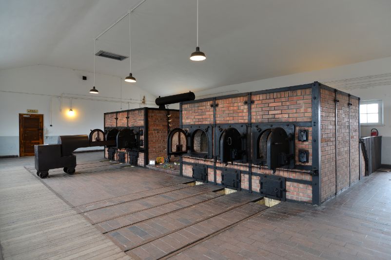 Six crematory ovens in an otherwise empty room. They are open. Small rails lead to the doors. In front of the oven on the far left, one of the rolling racks is still standing on the rails.