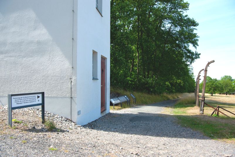 Ein Weg, der an Überesten des Lagerzauns entlangführt. Auf der linken Seite ein Wachturm. Davor steht ein Schild, das mitteilt, dass es sich bei dem Weg um den Postenweg handelt.