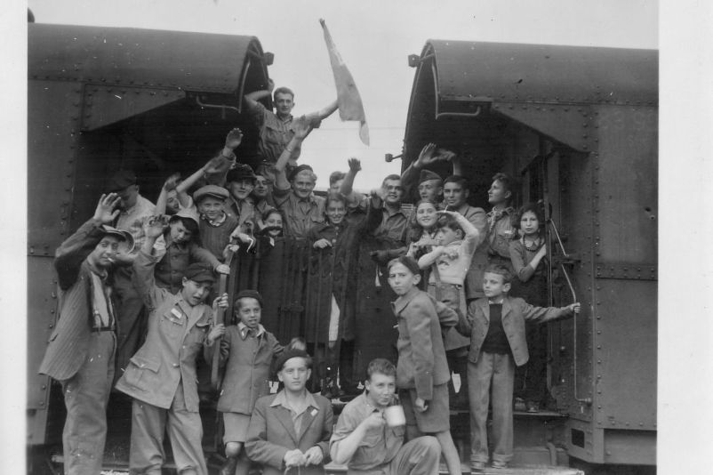 A group of children and young people stand by two railroad cars and wave at the camera.