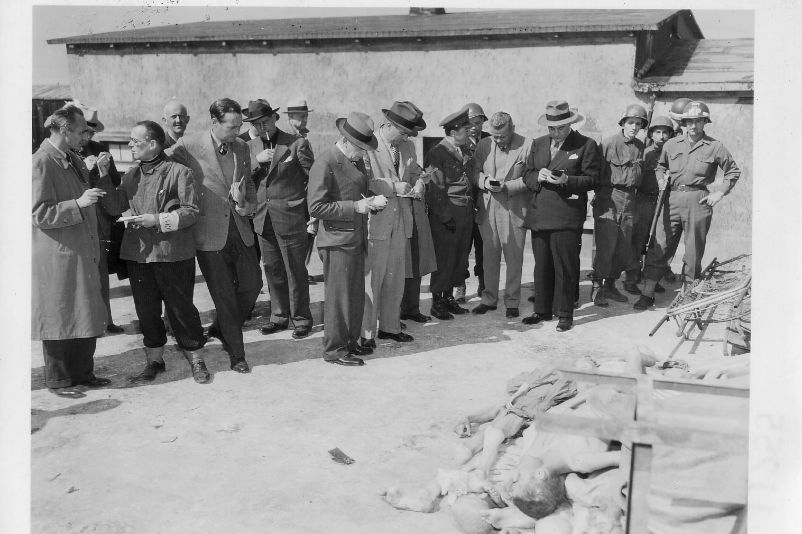 Ein Dutzend Männer stehen nah beieinander und inspizieren den Innenhof des Krematoriums, einige Männer notieren sich Notizen in ihre Blöcke.