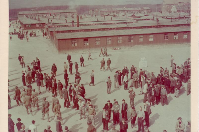 View from the gate building over the camp. Liberated prisoners and US soldiers are standing on the roll call square. The chamber building is in the far right background.