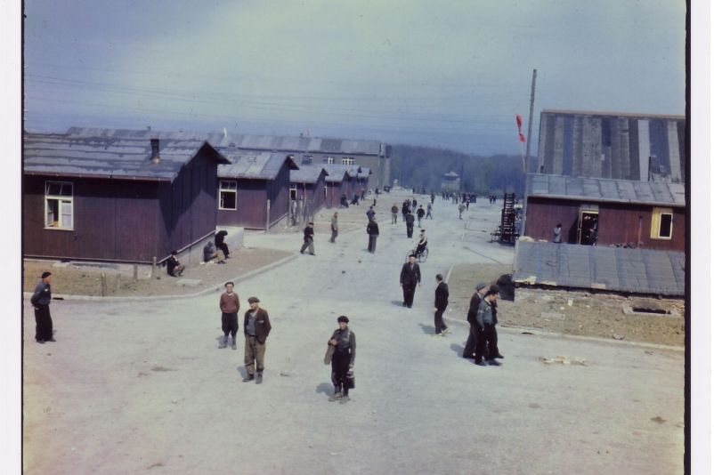 You can see liberated prisoners walking in small groups or individually along the camp street. To the right and left of the camp road, the one-story wooden prisoner barracks can be seen. On the left side, the stone barracks tower above them in the background.