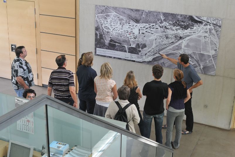 A group of visitors stands in front of a map, while an employee shows something on a bird's-eye view of the camp.