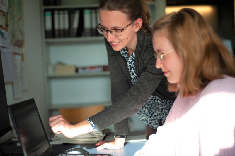 Eine Mitarbeiterin der KZ-Gedenkstätte Mittelbau-Dora zeigt einer jungen Freiwilligendienstleistenden etwas auf einem Laptop.