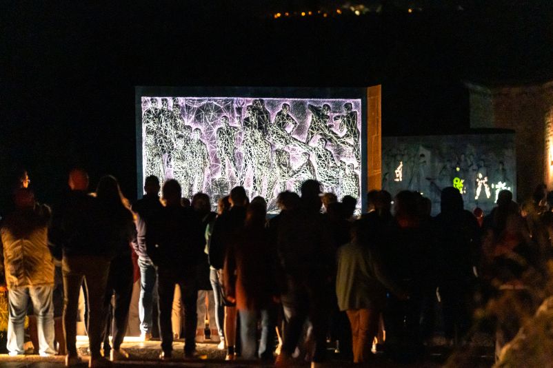 Menschenmenge betrachtet bei Nacht eine Videoprojektion auf einem Mahnmalrelief am Buchenwald-Denkmal, die stilisierte Figuren mit geometrischen Netzstrukturen zeigt; im Hintergrund sind weitere Lichtprojektionen auf Steinreliefs sichtbar.