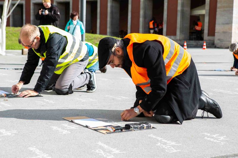 Two people in high-visibility vests kneel on the ground and write on the floor with chalk. 